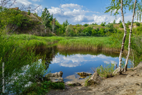 A lake surrounded by hills and forest. The sky is very brightly reflected in the calm water. There are stones, birches and reeds with sedge on the shore.