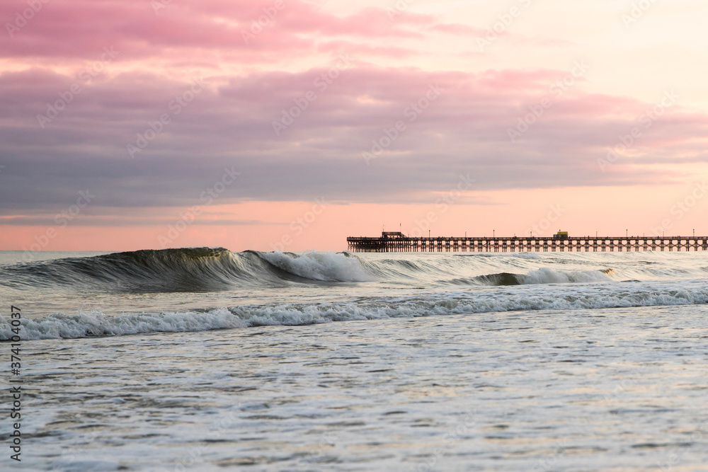 A wave rolling in under a pink and purple sky at sunset. The Apache Pier is visible on the horizon.