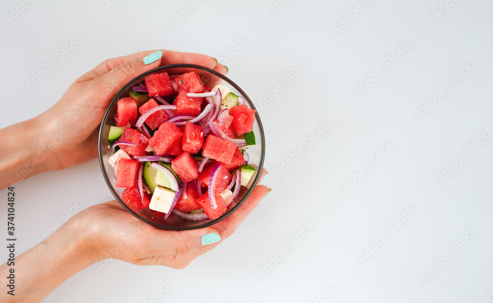 Woman's hands holding glass bowl with fresh delicious salad with watermelon, feta cheese, cucumber and red onions on white background. Close-up. Top view. Copy space. Healthy food concept. Woman's hands holding glass bowl with fresh delicious salad with watermelon, feta cheese, cucumber and red onions on white background. Close-up. Top view. Copy space. Healthy food concept.
