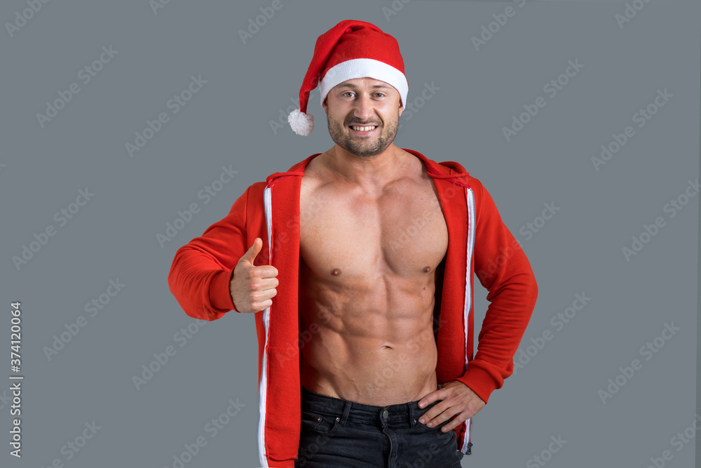 Handsome muscular man in santa claus hat showing thumbs up, isolated on gray background. Handsome muscular man in santa claus hat showing thumbs up, isolated on gray background.