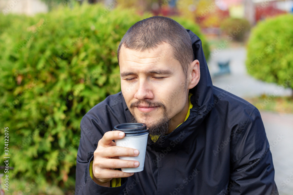 Handsome young man keeping eyes closed and smiling while smelling cup of fresh aromatic morning coffee. Outdoors in the park. Takeaway food. Handsome young man keeping eyes closed and smiling while smelling cup of fresh aromatic morning coffee. Outdoors in the park. Takeaway food.