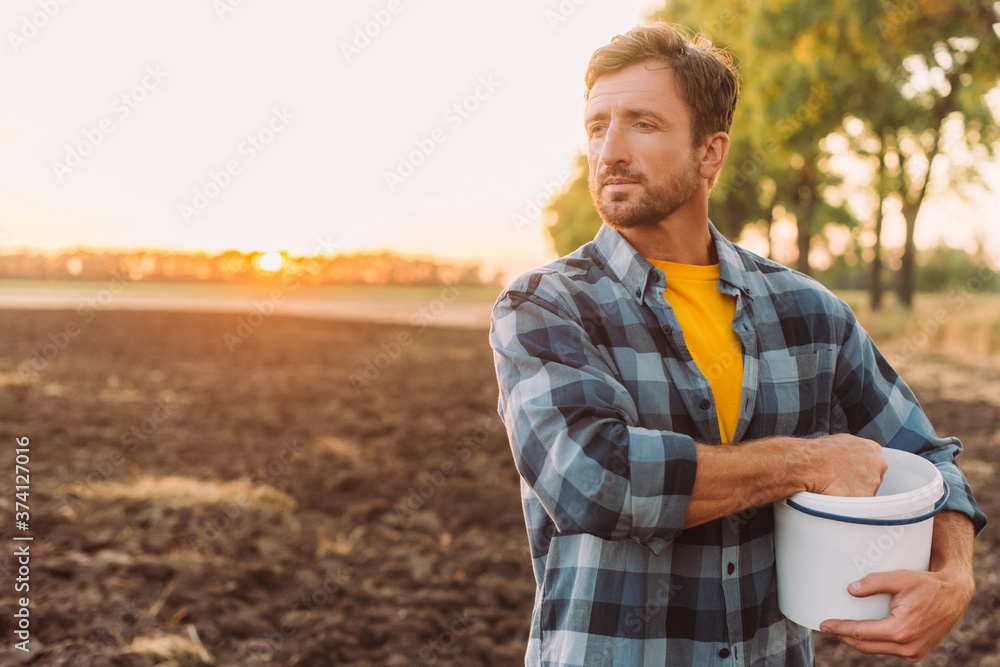 rancher in plaid shirt holding bucket while standing on plowed field in sunshine rancher in plaid shirt holding bucket while standing on plowed field in sunshine