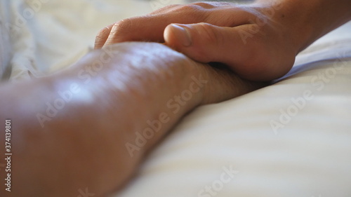 Grandson holds and comforts hand of his old grandmother in medical clinic. Young man gently touches wrinkled arm of sick mature grandma lying in bed hospital. Son shows care and love to his mother
