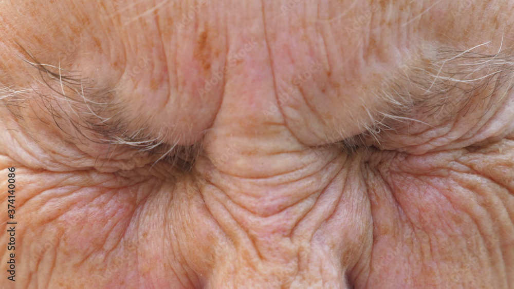 Fototapeta premium Portrait of mature grandmother looks into camera with amazed facial expression. Close up of elder grandma strongly screws up her gray eyes. Wrinkled mature female face shows surprise emotions