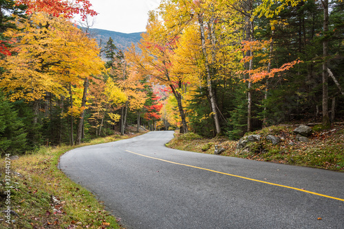 Fototapeta Naklejka Na Ścianę i Meble -  Steep winding  mountain road through a thick forest on a cloudy autumn day. Stunning autumn colours. Mount Washington, NH, USA.