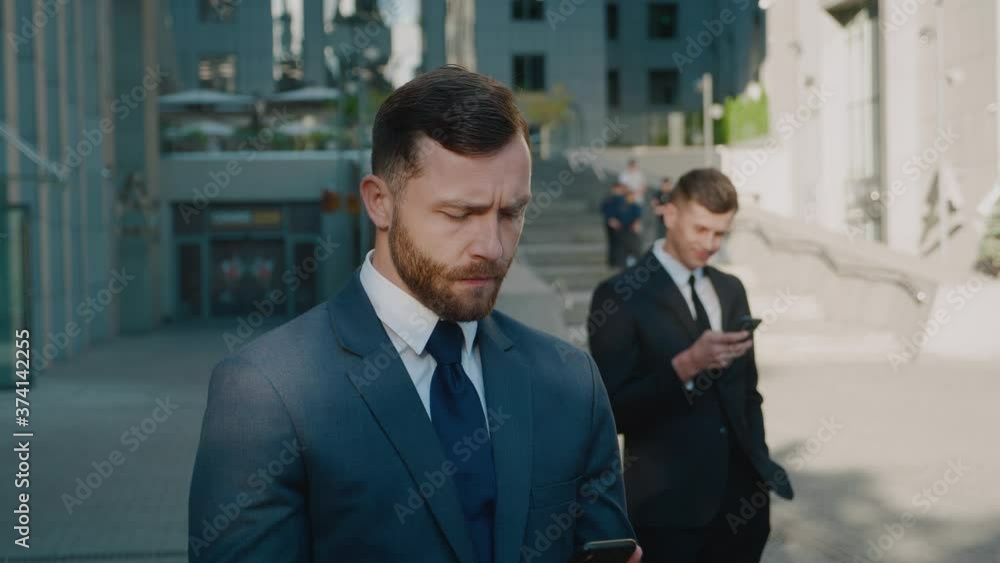 Caucasian young bearded man in official style using his smartphone near modern office building. Confident businessman using mobile app outdoors in business district downtown. Caucasian young bearded man in official style using his smartphone near modern office building. Confident businessman using mobile app outdoors in business district downtown.