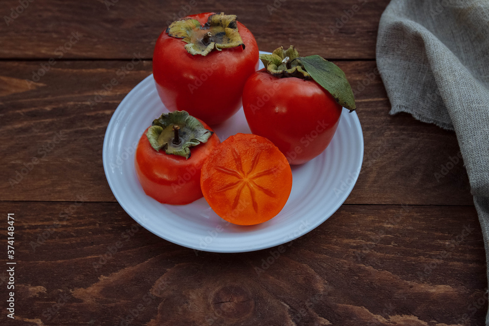 Red, ripe, juicy and soft persimmon on a white plate whole and cut in half with green leaves on a brown wooden background with a linen napkin, seasonal fruit concept, autumn harvest, organic food and Red, ripe, juicy and soft persimmon on a white plate whole and cut in half with green leaves on a brown wooden background with a linen napkin, seasonal fruit concept, autumn harvest, organic food and