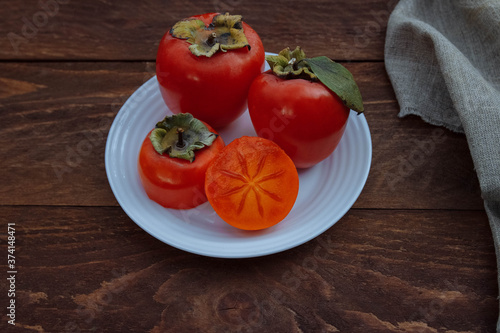 Red, ripe, juicy and soft persimmon on a white plate whole and cut in half with green leaves on a brown wooden background with a linen napkin, seasonal fruit concept, autumn harvest, organic food and 