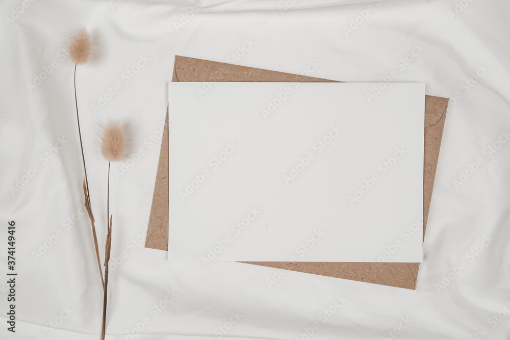 Fototapeta premium Blank white paper on brown paper envelope with Rabbit tail dry flower on white cloth. Mock-up of horizontal blank greeting card. Top view of Craft envelope on white background. Flat lay minimalism