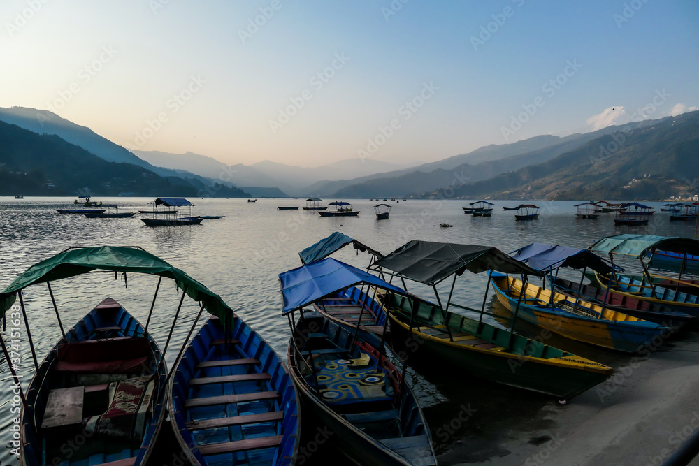 A view on Phewa Lake in Pokhara, Nepal with many colorful boats parked along it's shore. There are high Himalayan ranges in the back. Calm surface of the lake. Clear and sunny day. Serenity