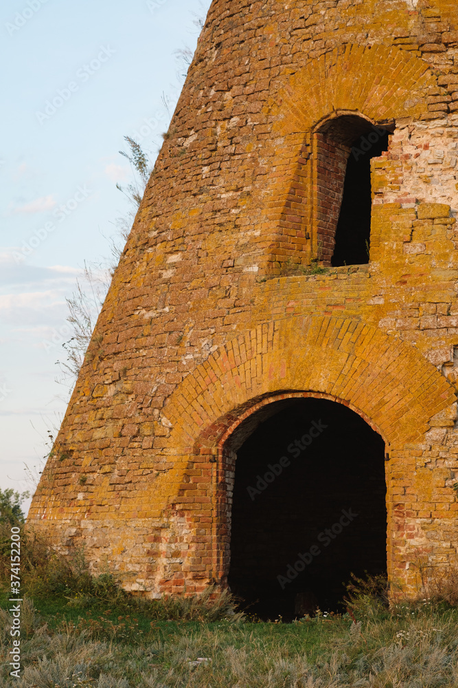 An old brick building stands alone in a field. An old dilapidated mill against the backdrop of a summer rural landscape. The concept of tourism, travel, exploring the history of the locality.