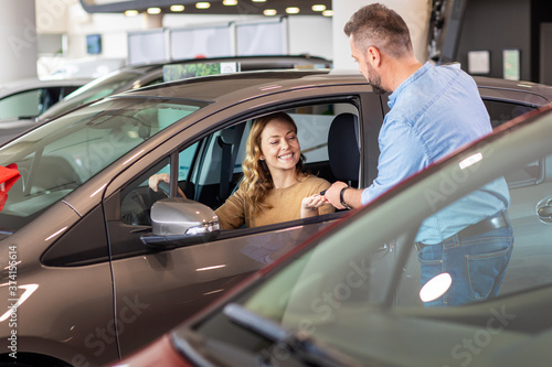 Smiling mid-adult couple buying a new car from dealership, woman sitting inside the vehicle while man standing beside her.