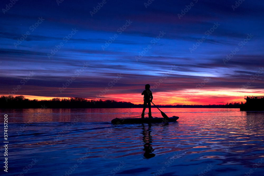 A little boy's silhouette on the SUP (stand up paddle) after sunset against a colorful sky at autumn or winter season