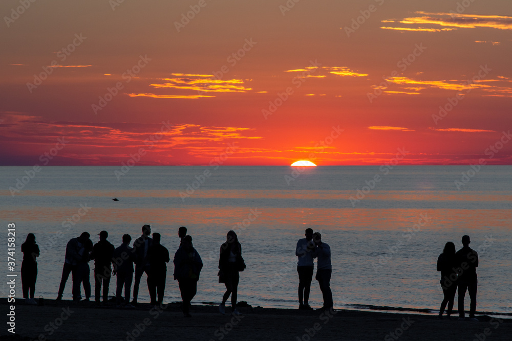Beautiful sunset at the sea. Young people walk along the beach. People are watching the sunset. Sea and red-yellow sky. Silhouettes of people on the background of the sunset. In the evening by the sea