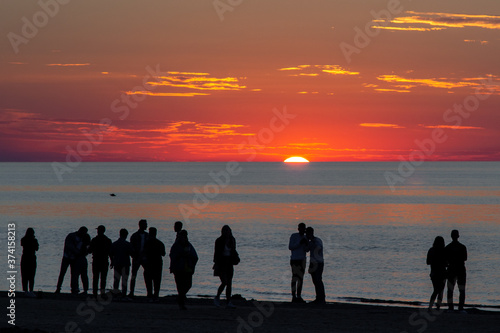 Beautiful sunset at the sea. Young people walk along the beach. People are watching the sunset. Sea and red-yellow sky. Silhouettes of people on the background of the sunset. In the evening by the sea