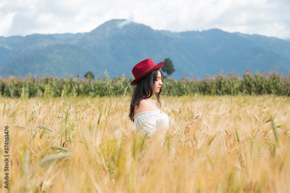 Young woman standing in the middle of a golden wheat field enjoying the sunset - girl with red hat in a field surrounded by mountains - woman alone in the field