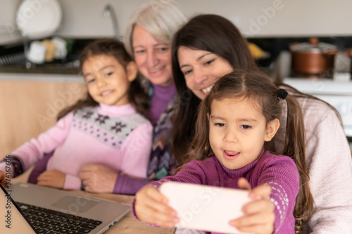Happy cheerful brazilian young child with smart cell phone taking photos of her family in kitchen home, indoors. Family, affectionate, reunion, together concept.