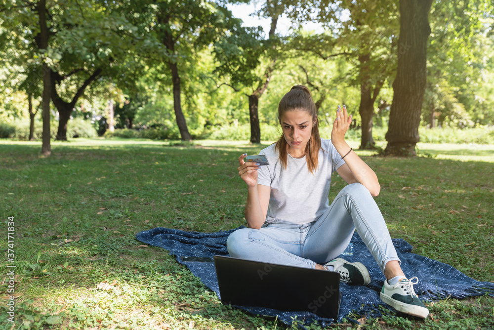 Young woman sitting in the park with laptop having issue with bank card and online shopping Young woman sitting in the park with laptop having issue with bank card and online shopping