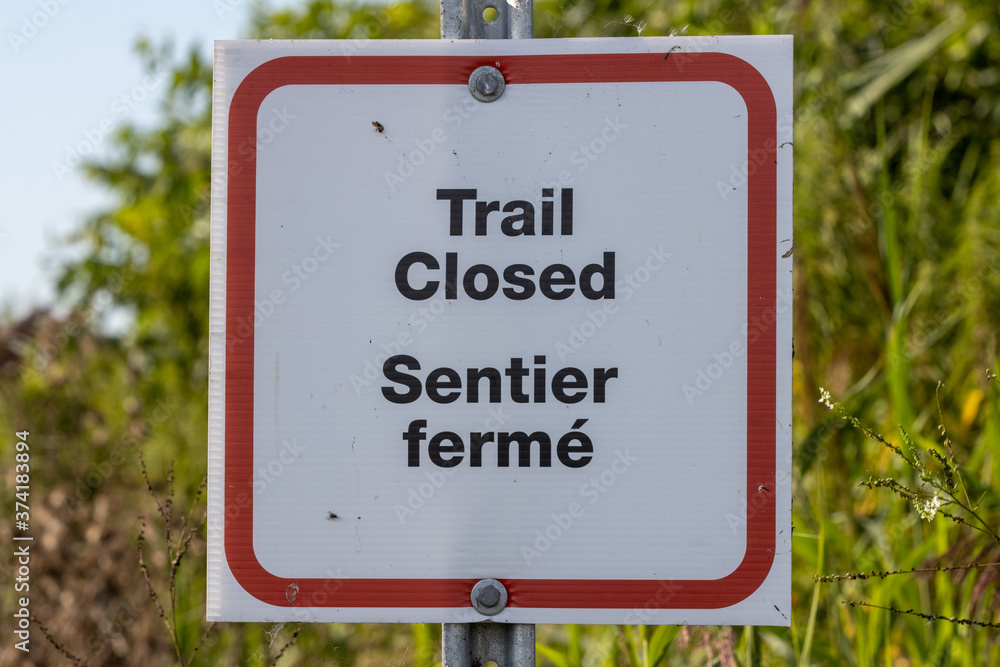 Centered Trail closed sign in English and French with overgrown trail in background