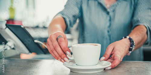 Closeup female barista hands holding cup of aromatic latte or cappuccino coffee in bar or cafe