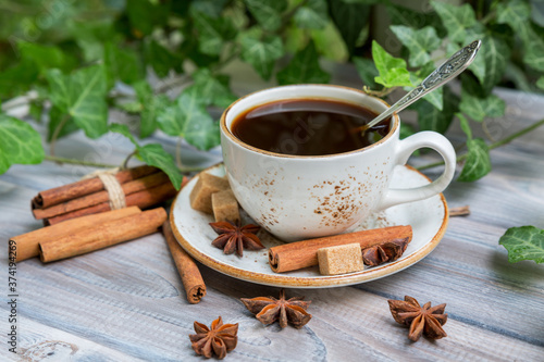 White cup of coffee with cinnamon and aniseed stars on a light wooden background with green ivy. Selective focus