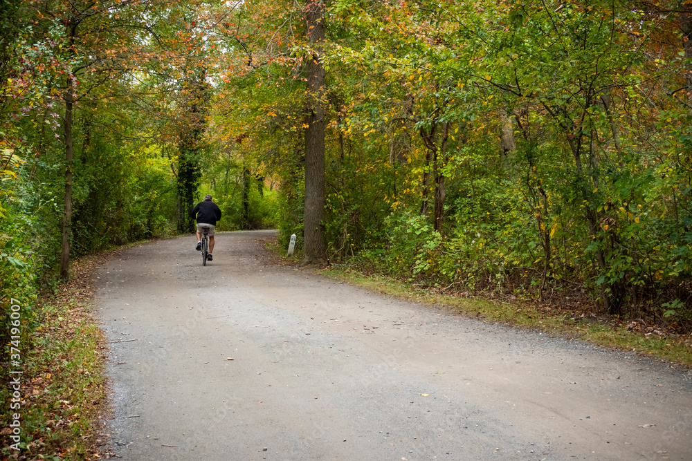 A senior man riding his bicycle down a gravel trail beneath colorful fall leaves in Belmont Lake State Park, Long Island, New York A senior man riding his bicycle down a gravel trail beneath colorful fall leaves in Belmont Lake State Park, Long Island, New York