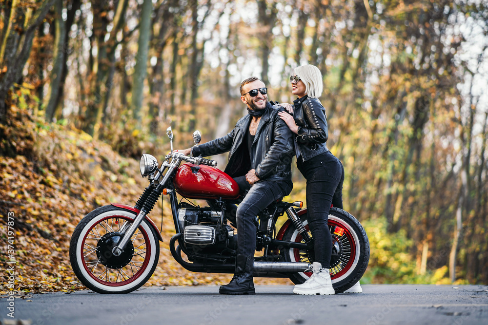 Fototapeta premium Pretty couple near red motorcycle on the road in the forest with colorful blured background