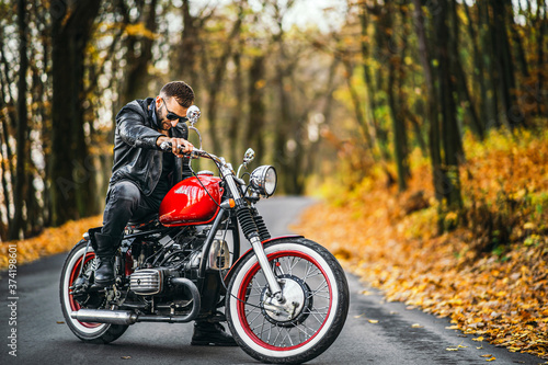 Bearded brutal man in sunglasses and leather jacket sitting on a motorcycle on the road in the forest © bedya