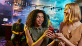Attractive young women looking at each other while toasting, posing with cocktail in their hands. Friends celebrating, having fun in the bar