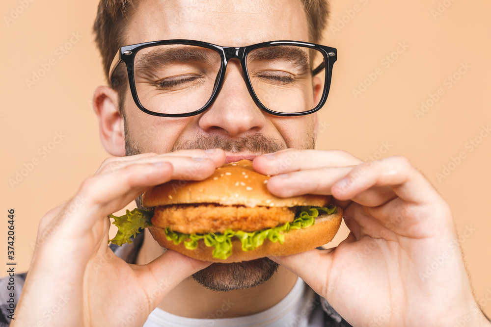 Young man holding a piece of hamburger. Student eats fast food. Burger is not helpful food. Very hungry guy. Diet concept. Isolated over beige background. Young man holding a piece of hamburger. Student eats fast food. Burger is not helpful food. Very hungry guy. Diet concept. Isolated over beige background.