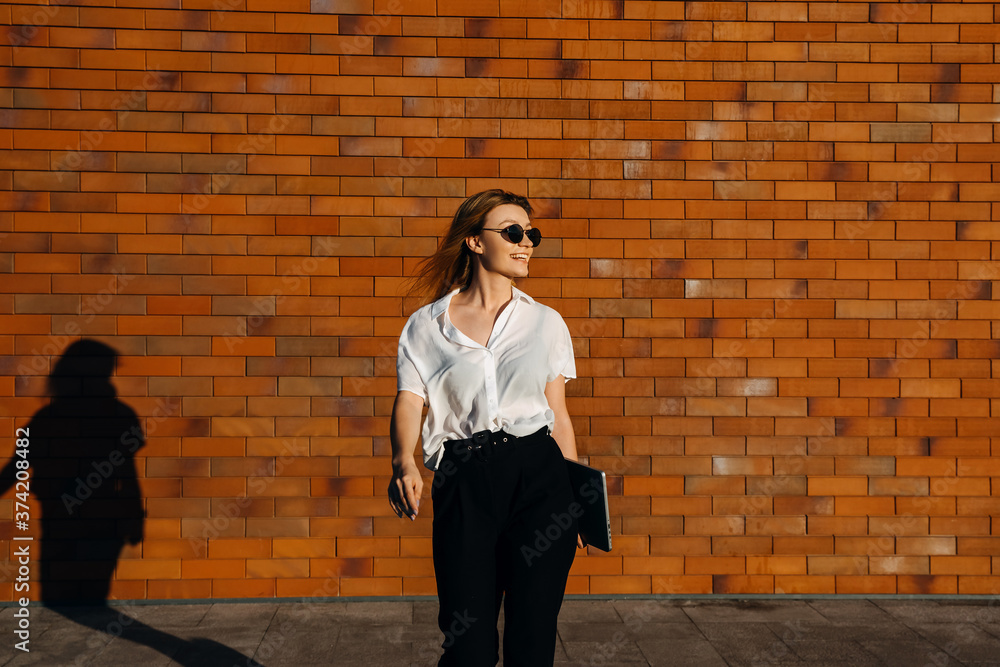 Young modern woman wearing white shirt and black pants, walking on the street, on a brick wall background, with a laptop, smiling.