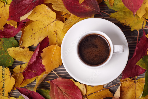Top view of white cup of coffee and autumn yellow leaves around on wooden table.