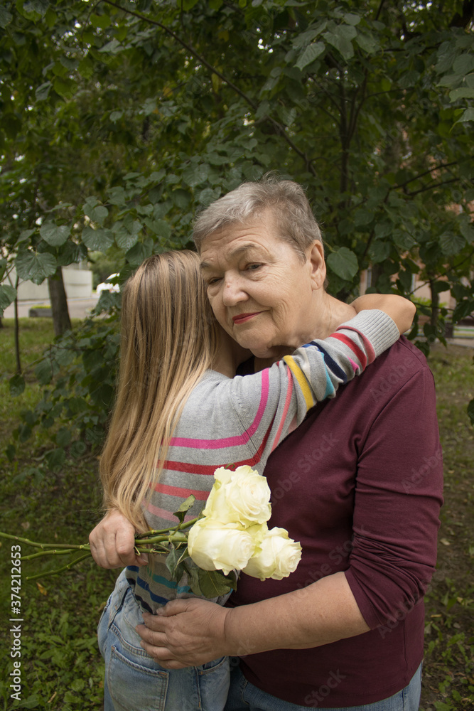 Grandmother hugs her granddaughter, who gave her flowers. The concept of love and friendship between generations. International Day of Older Persons and Grandparents Day Grandmother hugs her granddaughter, who gave her flowers. The concept of love and friendship between generations. International Day of Older Persons and Grandparents Day