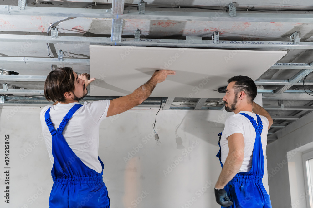 Obraz premium Two men are making a plasterboard ceiling. They use a drill and ladders