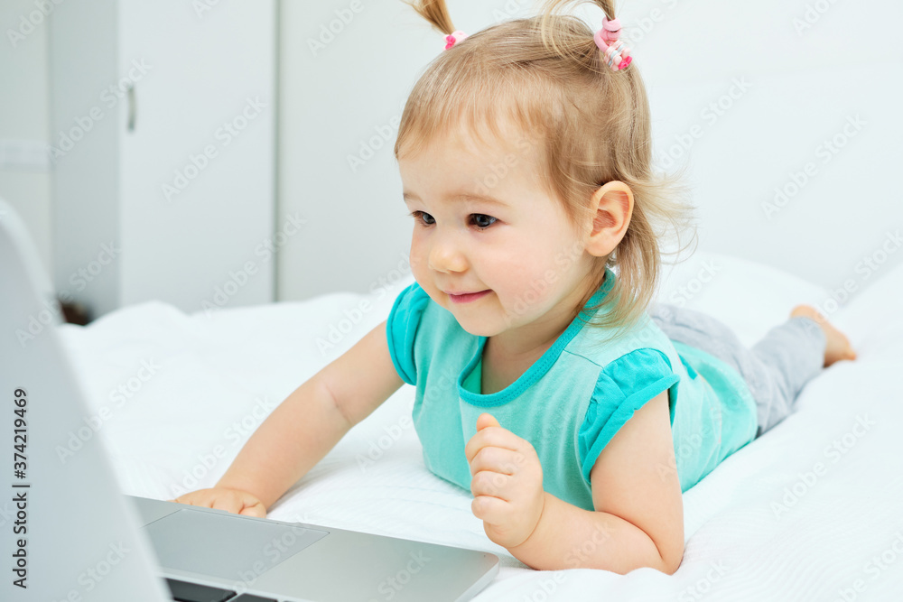 The child carefully looks into the laptop monitor while lying on the bed
