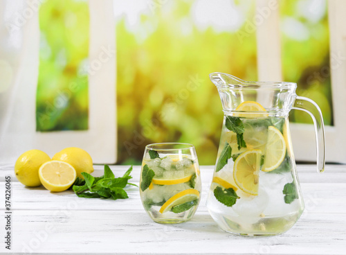 lemonade with lemon and mint. Lemonade with lemon and mint in a jug and in a glass stand diagonally on a wooden window sill against the background of an open window, close-up side view.