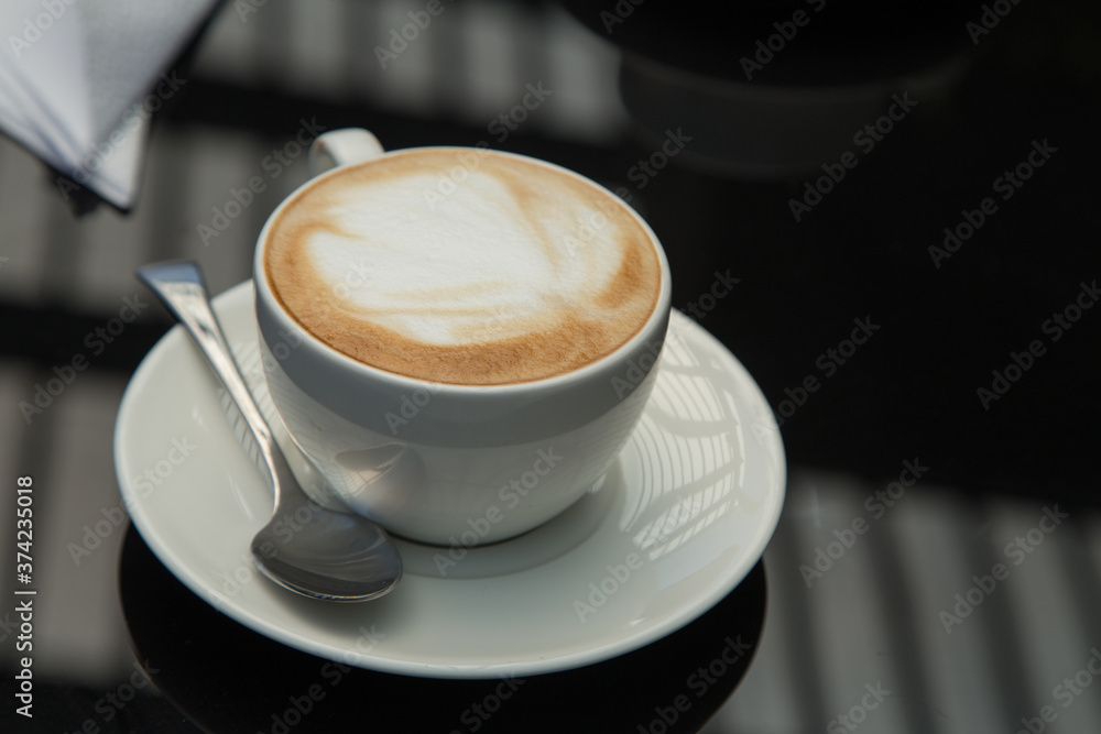 small white cup with a design in the coffee foam with a plate, spoon, napkin, on a smooth black surface where window lighting is reflected small white cup with a design in the coffee foam with a plate, spoon, napkin, on a smooth black surface where window lighting is reflected