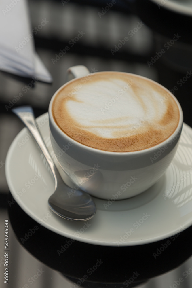 small white cup with a design in the coffee foam with a plate, spoon, napkin, on a smooth black surface where light from the window is reflected, close up shot