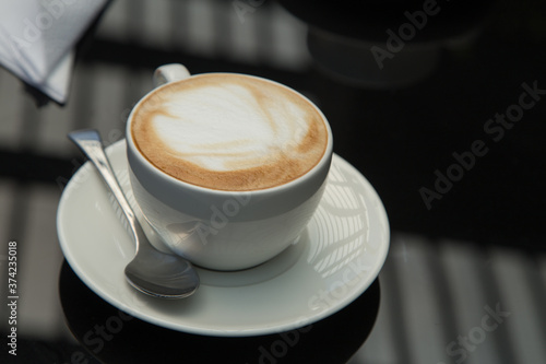 small white cup with a design in the coffee foam with a plate  spoon  napkin  on a smooth black surface where window lighting is reflected