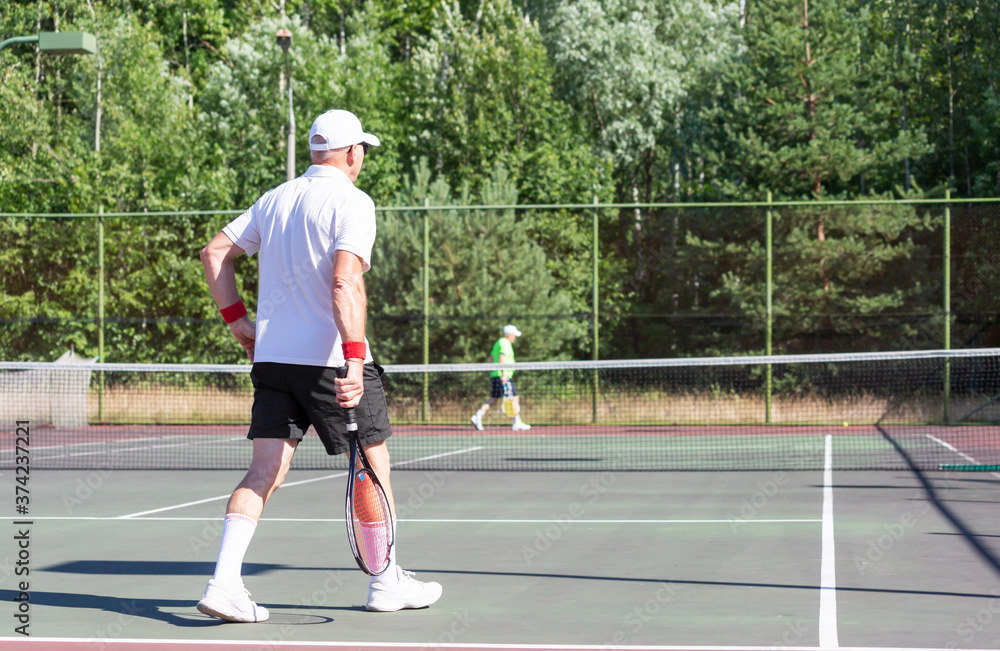 An elderly tennis player walks along the net on an outdoor court