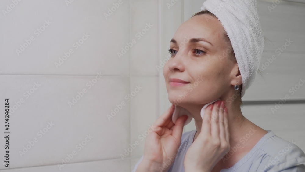 Adult woman with a towel on her head applying lotion and looking in mirror enjoy natural beauty and healthy Adult woman with a towel on her head applying lotion and looking in mirror enjoy natural beauty and healthy