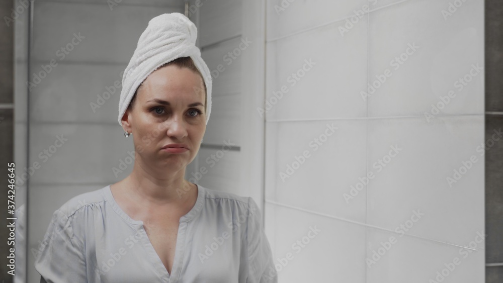 Frustrated Adult woman with a towel on her head looks at her face in the bathroom mirror Frustrated Adult woman with a towel on her head looks at her face in the bathroom mirror