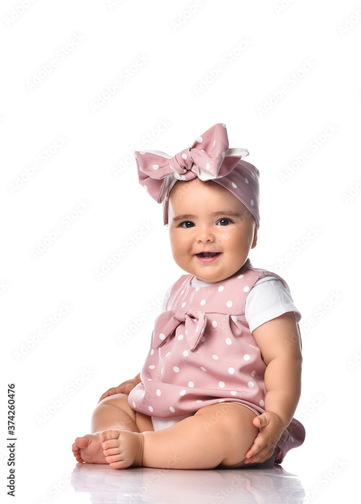 Happy laughing infant baby toddler in polka dot dress and headband with bow sits on the floor holding hand at her leg Happy laughing infant baby toddler in polka dot dress and headband with bow sits on the floor holding hand at her leg