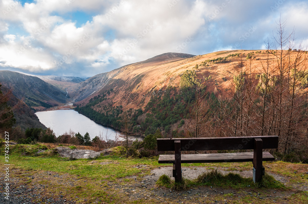 View over Upper Lake in Glendalough Scenic Park, a lot of hills, sun and clouds, bench on the front. Concepts: landscape, outdoor, tourism, travel