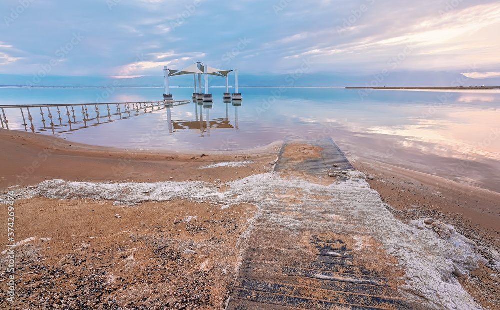 Calm morning at Ein Bokek Dead Sea beach, pink clouds reflection over water surface near steel rails leading to sun shade shelter, sand covered with salt crystals