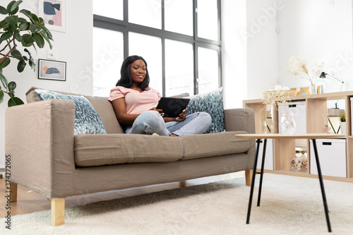 people, technology and leisure concept - happy young african american woman sitting on sofa with tablet pc computer at home