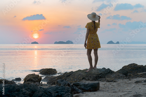 Young caucasian woman in hat and yellow dress on the sandy beach enjoying the sunset