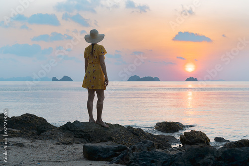 Young caucasian woman in hat and yellow dress on the sandy beach enjoying the sunset