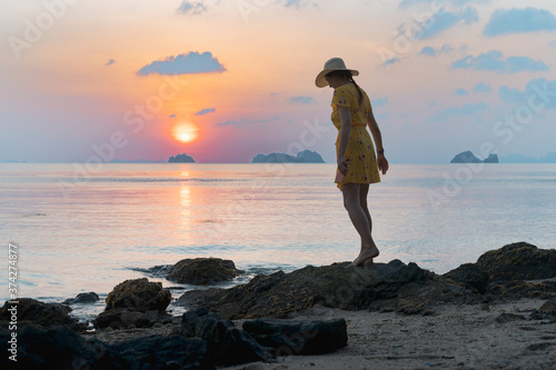 Young caucasian woman in hat and yellow dress walks on the sandy beach and enjoying the sunset