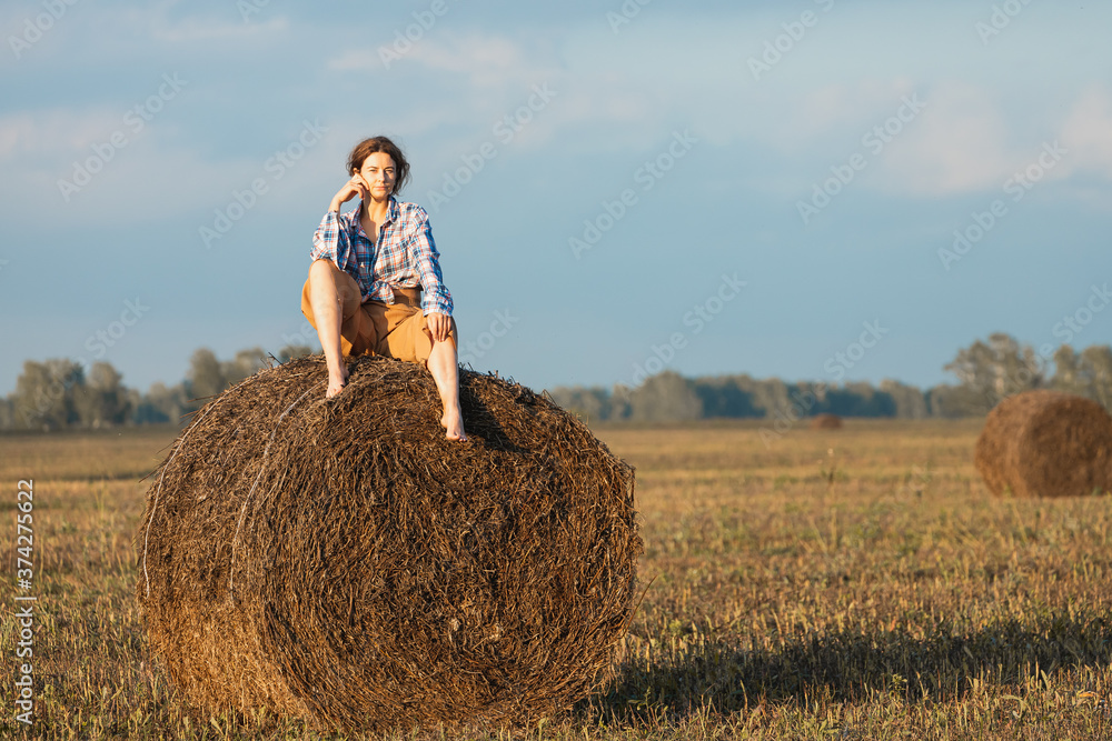 Beautiful modern  young woman wearing in beige trousers and a plaid shirt posing against   a haystack in field. Trendy teenage girl in autumn outdoors.